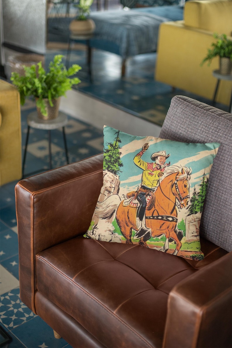 May include: A brown leather couch with a throw pillow featuring a vintage illustration of a cowboy riding a horse through a forest. The pillow has a light blue, green, and yellow color scheme.