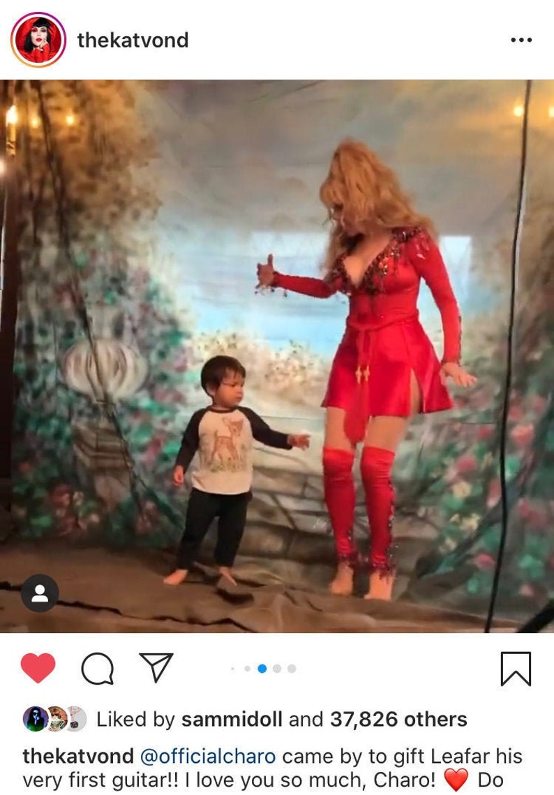 May include: A woman in a red sequined dress and red leggings is standing in front of a painted backdrop. She is holding her hand up in the air, as if she is dancing. A young boy in a grey and white shirt and black pants is standing in front of her, looking at her. The text "thekatvond @officialcharo came by to gift Leafar his very first guitar!! I love you so much, Charo! Do" is written below the image.