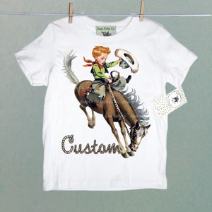 K&ouml;nnte beinhalten: Wei&szlig;es T-Shirt mit einem Grafikdruck eines Jungen, der ein sich aufb&auml;umendes Pferd reitet. Der Junge tr&auml;gt einen Cowboyhut und ein rotes Bandana. Der Text "Custom" ist unter dem Bild gedruckt.