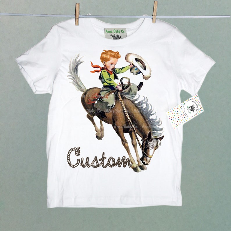 K&ouml;nnte beinhalten: Wei&szlig;es T-Shirt mit einem Grafikdruck eines Jungen, der ein sich aufb&auml;umendes Pferd reitet. Der Junge tr&auml;gt einen Cowboyhut und ein rotes Bandana. Der Text "Custom" ist unter dem Bild gedruckt.