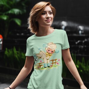 Peut inclure: Un t-shirt vert clair avec une illustration r&eacute;tro d'un agneau et d'un enfant. Le graphisme repr&eacute;sente l'agneau et l'enfant pr&egrave;s d'une cl&ocirc;ture en bois avec des fleurs. Le t-shirt est fait d'une mati&egrave;re douce.