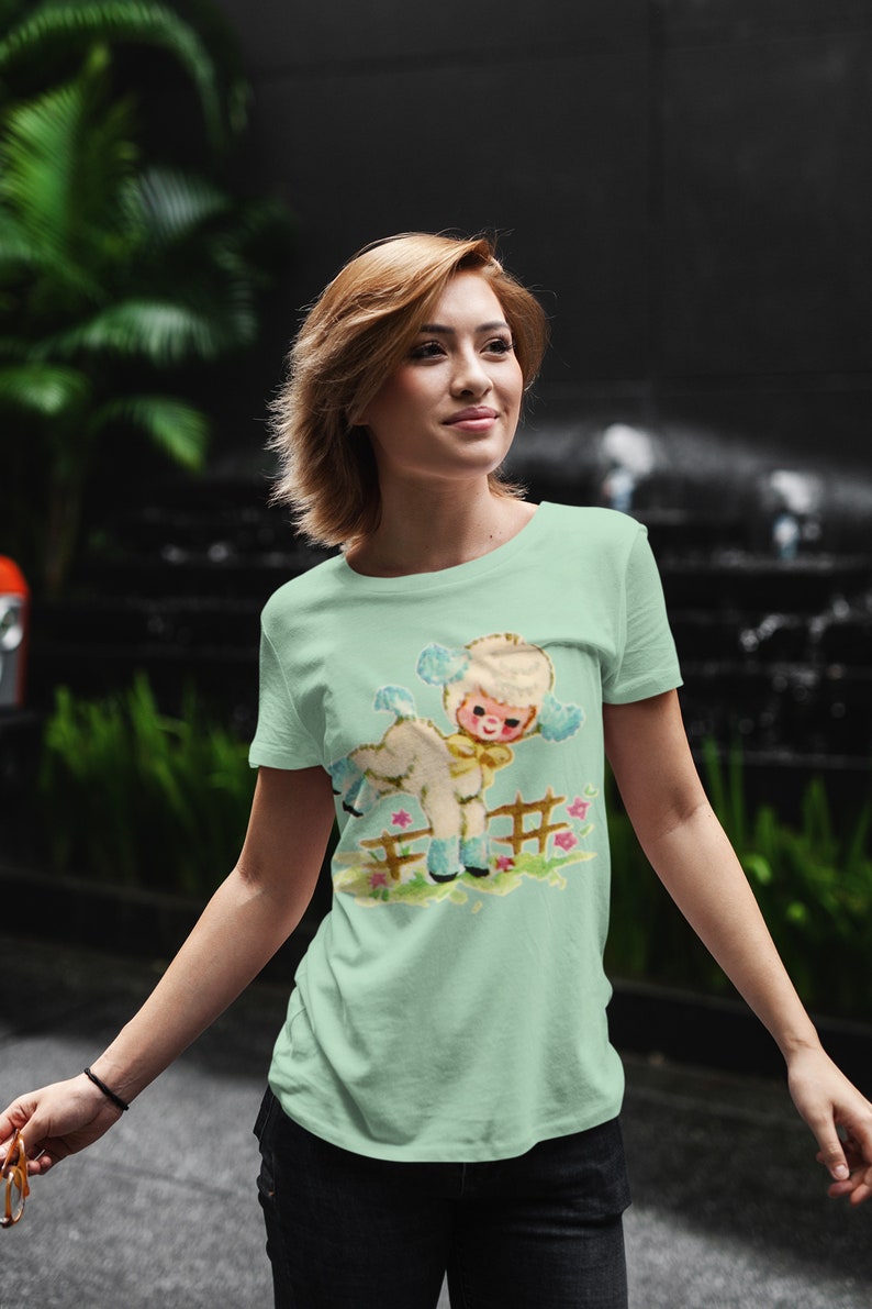 Peut inclure: Un t-shirt vert clair avec une illustration r&eacute;tro d'un agneau et d'un enfant. Le graphisme repr&eacute;sente l'agneau et l'enfant pr&egrave;s d'une cl&ocirc;ture en bois avec des fleurs. Le t-shirt est fait d'une mati&egrave;re douce.