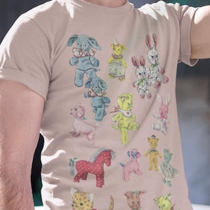 Puede incluir: Una camiseta rosa claro con un estampado vintage de varios juguetes de peluche. Los juguetes incluyen un caballo, un gato, un perro, un conejo y un oso. Los juguetes son de diferentes colores y patrones.