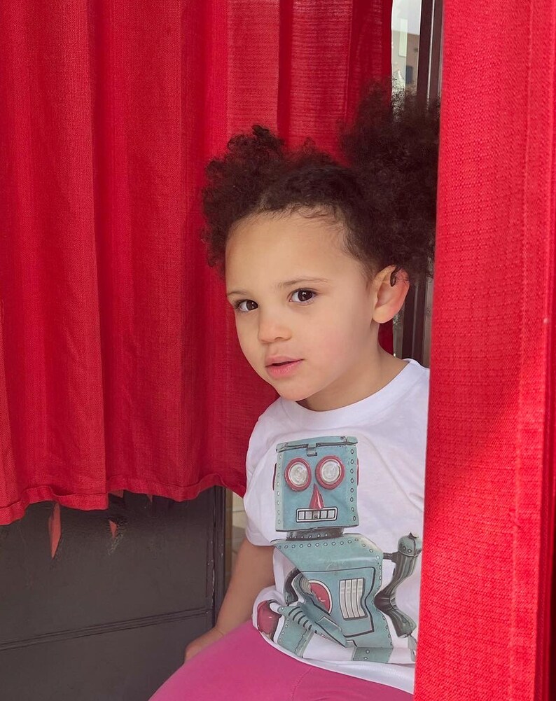 Puede incluir: Una ni&ntilde;a con el pelo casta&ntilde;o rizado lleva una camiseta blanca con un robot vintage. El robot es azul y gris con ojos rojos y una nariz roja. La ni&ntilde;a lleva pantalones rosas y asoma por detr&aacute;s de una cortina roja.