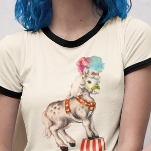 May include: A white t-shirt with a black ringer neckline features a vintage illustration of a white pony wearing a colorful hat and standing on a red and white striped circus platform. The text "CIRCUS" is printed below the pony in rainbow colors.