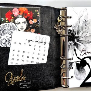 Może przedstawiać: Czarny skórzany planer z metalowym segregatorze w kolorze złotym. Planer zawiera kalendarz na styczeń, ozdobną kartę z portretem kobiety i kwiatowym wzorem. Złoty charms z kwiatami i perłowymi akcentami wisi na segregatorze.