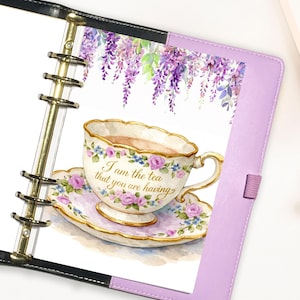 May include: A planner with a watercolor illustration of a teacup and saucer, adorned with pink roses and blue flowers, and the text "I am the tea that you are having." The background features purple wisteria. The planner has a lavender cover and gold accents.