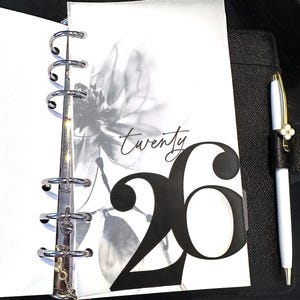 以下が含まれることがあります： ページに白黒の花柄デザインが施された、開いた状態の手帳。ページには筆記体で「twenty」の文字と、太字の数字「26」が表示されています。右側には花のチャームが付いた白いペンがあります。