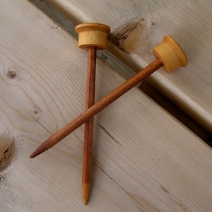 Może przedstawiać: Dwie drewniane druty do rob&oacute;tek ręcznych z zaokrąglonymi końc&oacute;wkami i naturalnym wykończeniem z drewna. Druty są skrzyżowane i leżą na jasnym drewnianym podłożu.