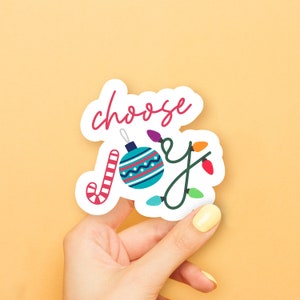 May include: White sticker with the text "choose joy" in pink script. The sticker features a blue and white Christmas ornament with a red candy cane and a string of colorful lights.