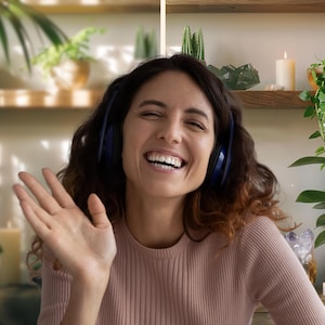 May include: A woman with curly brown hair is wearing blue headphones and a pink sweater. She is smiling and waving with her right hand. The background is a shelf with plants, candles, and crystals.
