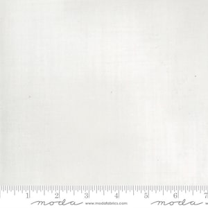 May include: A white fabric with a ruler along the bottom edge. The fabric has a subtle texture and appears to be cotton or a similar material. The ruler is marked in inches, with the "moda" logo and website address printed below.