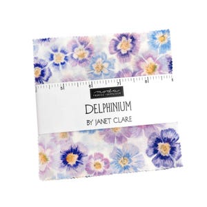 Op de afbeelding: Een stapel vierkante stukken stof met een bloemenpatroon in tinten paars, blauw en wit. Een witte papieren band wikkelt zich om het midden, met de tekst "MODA FABRICS + SUPPLIES", "DELPHINIUM" en "BY JANET CLARE" en een liniaal van 5 tot 9 inch.