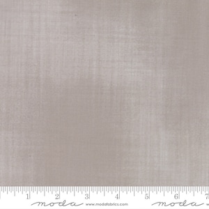 May include: A piece of fabric in a solid, muted taupe color. The fabric has a subtle texture and a ruler along the bottom edge, marked in inches. The fabric is from Moda Fabrics.