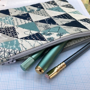 May include: A zippered pouch with a geometric pattern in shades of blue, gray, and green. The pouch is open and contains two green pens and a black pencil with a gold tip.