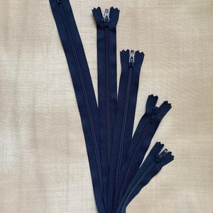 May include: Five black zipper closures of varying lengths. The zippers are arranged in a fan shape, with the longest zipper at the top and the shortest zipper at the bottom.
