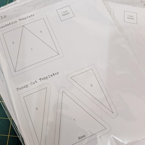 May include: A set of white paper foundation templates for quilting, labeled Iris, featuring numbered geometric shapes, triangles, and a 1-inch square reference guide. The sheets are enclosed in clear plastic sleeves, displaying Fussy Cut Templates.