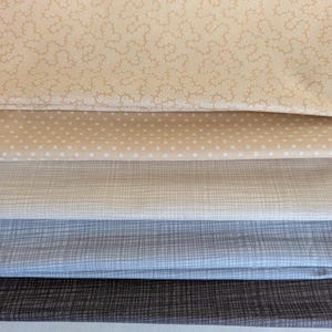 Pode incluir: Uma pilha de amostras de tecido em várias cores neutras. A camada superior é bege claro com um padrão floral repetido. Abaixo estão camadas de tecido em creme com bolinhas brancas, bege claro com um padrão de grade, azul claro com um padrão de grade e marrom escuro com um padrão de grade.