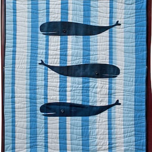 May include: A quilted wall hanging with three dark blue whale shapes on a blue and white striped background. The whales have button eyes and are set on a wavy pattern. The quilt is framed in a dark brown border.