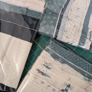 Könnte beinhalten: Mehrere Stoffpakete in transparenter Kunststoffverpackung. Die Stoffe weisen nautische Designs auf, darunter Segelboote, Leuchttürme und Sternmuster, in Blau-, Creme- und Schwarztönen. Die Pakete sind auf einer grünen Schneidematte angeordnet.