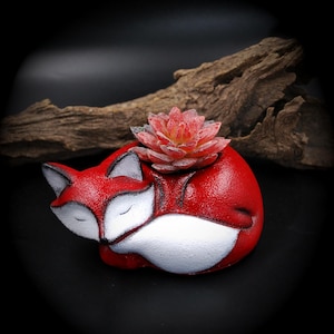 May include: A red fox-shaped planter with white accents and a succulent plant. The fox is curled up, appearing to be sleeping. The planter is a decorative item for small plants, adding a whimsical touch to any space.