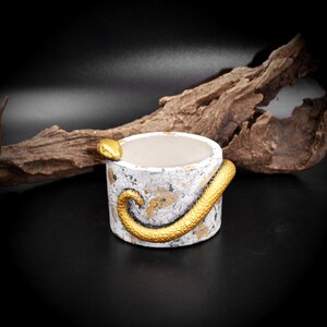 May include: A small, round, white and gold planter with a textured finish. A gold snake winds around the exterior. The interior is a light beige color. The planter is set against a dark background with a piece of driftwood.