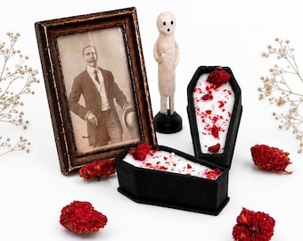 Può includere: Due candele nere a forma di bara con cera bianca cosparsa di petali di fiori secchi rossi, una piccola statuetta di fantasma color bianco sporco su base nera e una cornice in legno marrone scuro con un ritratto in bianco e nero di un uomo.