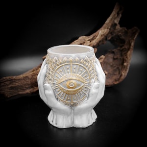 Puede incluir: Un recipiente decorativo blanco y dorado con un símbolo de ojo, sostenido por dos manos. El recipiente tiene forma cilíndrica con la parte superior abierta. El símbolo del ojo está en el centro, rodeado de líneas radiales. El fondo es oscuro.