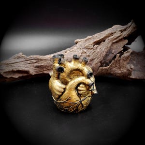 May include: A golden-colored anatomical heart sculpture with black accents. The heart's intricate details are visible, including veins and chambers. The sculpture is set against a dark background with a piece of driftwood.