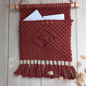 May include: A red macrame wall hanging with a diamond-shaped design and a pocket for holding mail or other items. The hanging has a wooden dowel at the top and a fringe of red and cream yarn at the bottom.