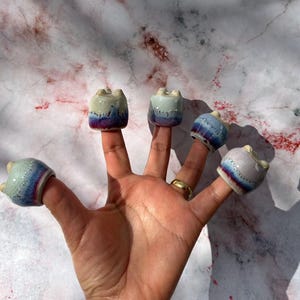 May include: Five small ceramic pots with a white, blue and purple glaze are worn on the fingers of a hand. The pots are all different shapes and sizes.