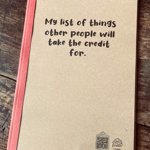 May include: A notebook with a tan cover and a coral spine. The cover has the text "My list of things other people will take the credit for." A QR code and the words "Jen's Ponder" are at the bottom of the page.