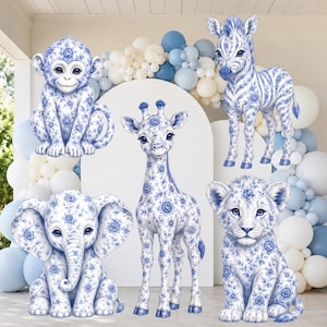 May include: A collection of five animal figures including a monkey, zebra, giraffe, elephant, and lion cub, all featuring a blue and white floral porcelain-style pattern. The animals are arranged in front of a white arch and balloon garland.