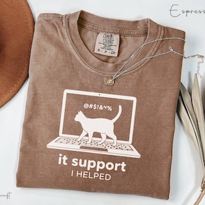 Op de afbeelding: Espresso-kleurig T-shirt met een witte afbeelding van een kat op een laptopscherm, met de tekst "it support I HELPED". Het shirt heeft een ronde hals en een Comfort Colors-label. Een zilveren ketting met een hanger ligt op het shirt.