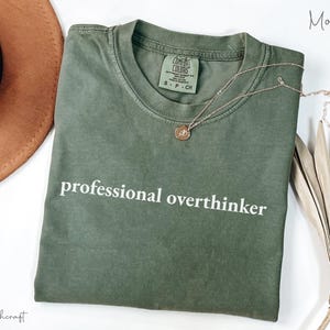 K&ouml;nnte beinhalten: Ein moosgr&uuml;nes Comfort Colors T-Shirt mit dem wei&szlig;en Aufdruck "professional overthinker". Eine goldene Halskette mit einem kleinen Anh&auml;nger liegt auf dem Shirt. Ein brauner Hut und eine getrocknete Pflanze befinden sich im Hintergrund.