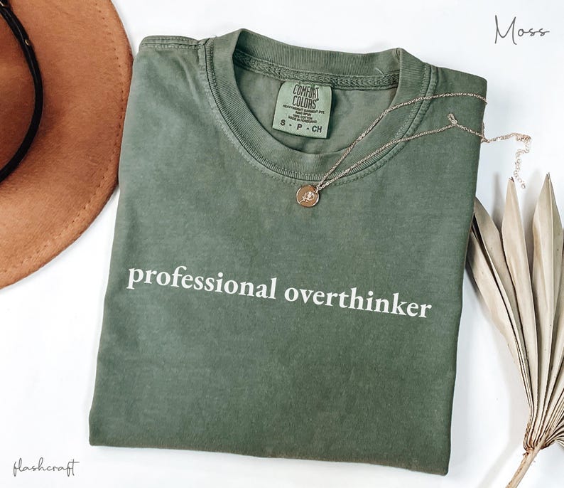 K&ouml;nnte beinhalten: Ein moosgr&uuml;nes Comfort Colors T-Shirt mit dem wei&szlig;en Aufdruck "professional overthinker". Eine goldene Halskette mit einem kleinen Anh&auml;nger liegt auf dem Shirt. Ein brauner Hut und eine getrocknete Pflanze befinden sich im Hintergrund.