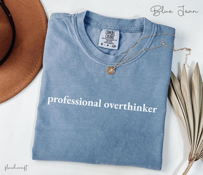 K&ouml;nnte beinhalten: Ein blaues Comfort Colors T-Shirt mit dem wei&szlig;en Aufdruck "professional overthinker". Eine goldene Halskette mit Anh&auml;nger liegt &uuml;ber dem Shirt. Das Shirt ist gefaltet und auf einer wei&szlig;en Oberfl&auml;che platziert.