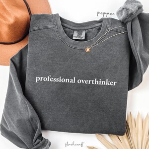 K&ouml;nnte beinhalten: Ein dunkelgraues Sweatshirt mit dem wei&szlig;en Text "professional overthinker". Das lang&auml;rmlige Oberteil hat einen Rundhalsausschnitt und ist aus einem weichen Material gefertigt. Eine goldene Halskette und ein brauner Hut sind ebenfalls sichtbar.