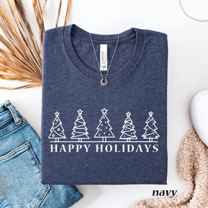 May include: Navy blue t-shirt with a white graphic of six Christmas trees and the text "HAPPY HOLIDAYS". The shirt is folded and displayed with blue jeans, white shoes, and a necklace.