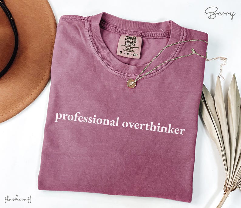 K&ouml;nnte beinhalten: Beerenfarbenes T-Shirt mit dem wei&szlig;en Text "professional overthinker". Das Shirt hat einen Rundhalsausschnitt und ein kleines Etikett auf der oberen Brust. Eine goldene Halskette mit Anh&auml;nger ist &uuml;ber dem Shirt drapiert. Ein brauner Hut ist oben links zu sehen.
