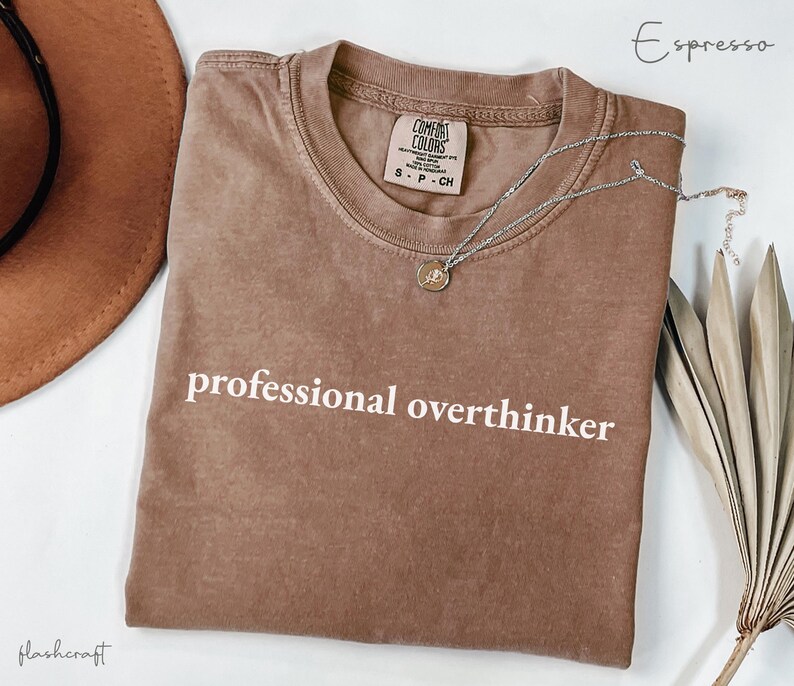 K&ouml;nnte beinhalten: Espressofarbenes T-Shirt mit dem wei&szlig;en Schriftzug "professional overthinker". Eine silberne Halskette mit kleinem Anh&auml;nger liegt auf dem Shirt. Das Shirt ist gefaltet, und ein brauner Hut ist teilweise sichtbar.