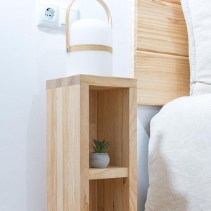 Op de afbeelding: Een lichtgekleurd houten nachtkastje met een kleine plank met een potplant. Bovenop staat een witte lamp met een houten handvat. Het nachtkastje staat naast een bed met een wit dekbed.