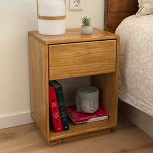 May include: A light brown wooden nightstand with a single drawer and an open lower shelf. On top sits a white portable lamp with a light wood handle and a small succulent in a gray pot. The lower shelf holds two books and a gray Buddha head statue.