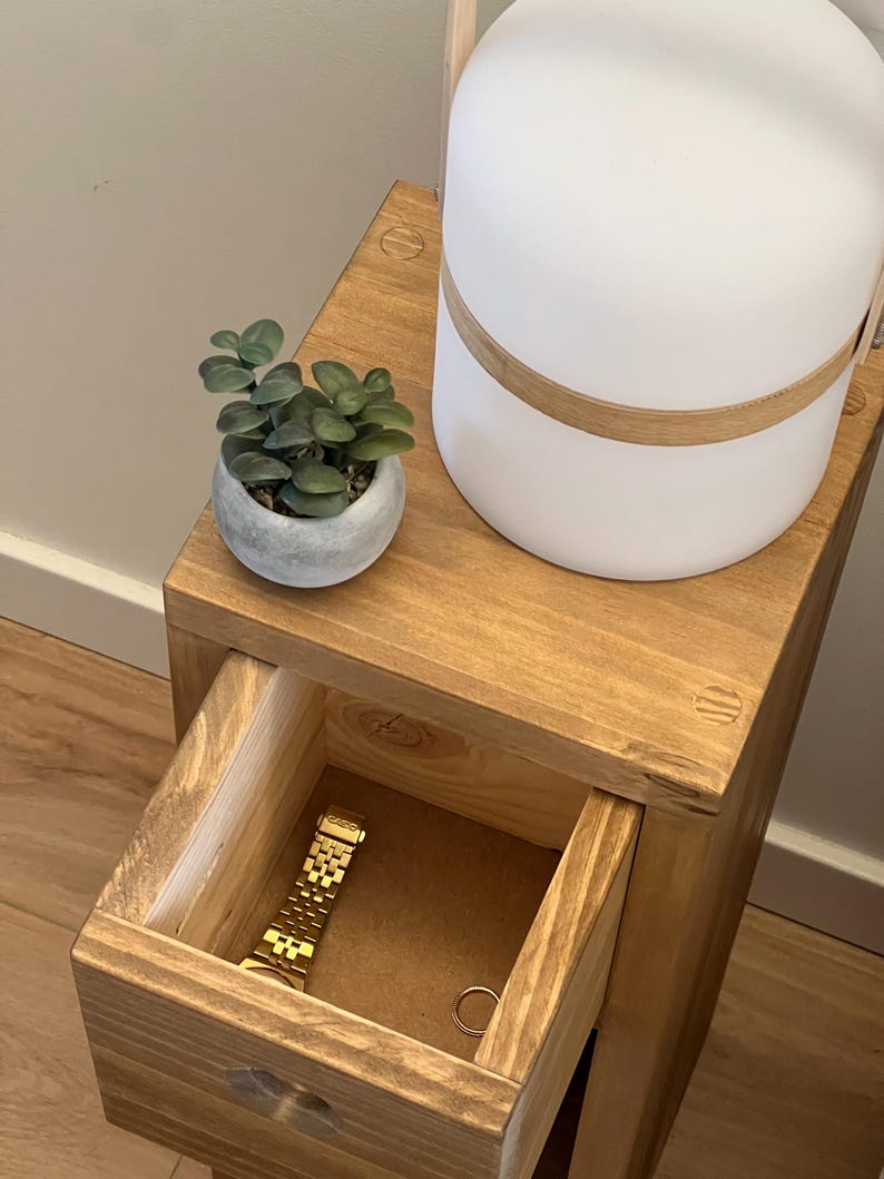 May include: A wooden bedside table with a drawer that is open, revealing a gold watch and a ring. A small potted succulent plant sits on the top shelf, next to a white lamp with a wooden base.