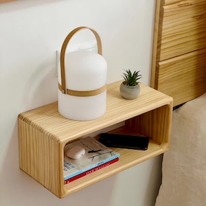 May include: A light-coloured wooden floating bedside table with a rounded design. It holds a white lamp with a wooden handle, a small potted plant, a book, a phone, and wireless earbuds. The table is mounted on a white wall.