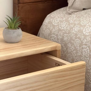 May include: A light-colored wooden nightstand with a single open drawer featuring the partial text BORN visible on the interior side. A small, round, gray concrete-style planter containing a green artificial succulent sits on the top surface.