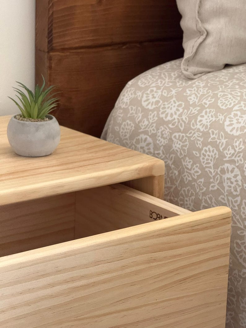 May include: A light-colored wooden nightstand with a single open drawer featuring the partial text BORN visible on the interior side. A small, round, gray concrete-style planter containing a green artificial succulent sits on the top surface.