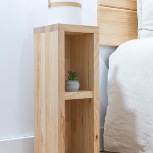 K&ouml;nnte beinhalten: Ein hoher, schmaler Nachttisch aus hellem Holz mit einem rechteckigen Rahmen und einem einzelnen horizontalen Regalboden in der Mitte. Das M&ouml;belst&uuml;ck besteht aus vertikalen Holzplanken und tr&auml;gt eine kleine, gr&uuml;ne Sukkulente im Topf.