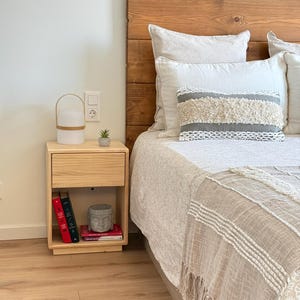 May include: A light-colored wooden nightstand with a single drawer and an open shelf containing three books and a small gray Buddha head statue. The nightstand is positioned next to a bed with a wooden headboard and textured bedding.