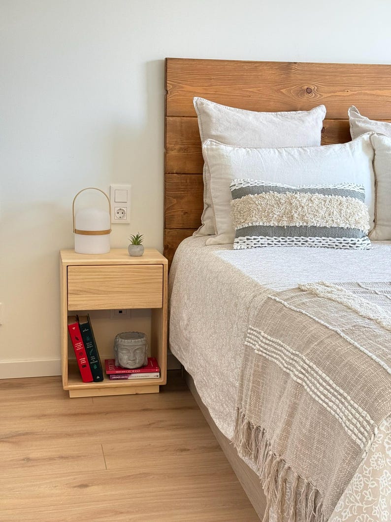 May include: A light-colored wooden nightstand with a single drawer and an open shelf containing three books and a small gray Buddha head statue. The nightstand is positioned next to a bed with a wooden headboard and textured bedding.
